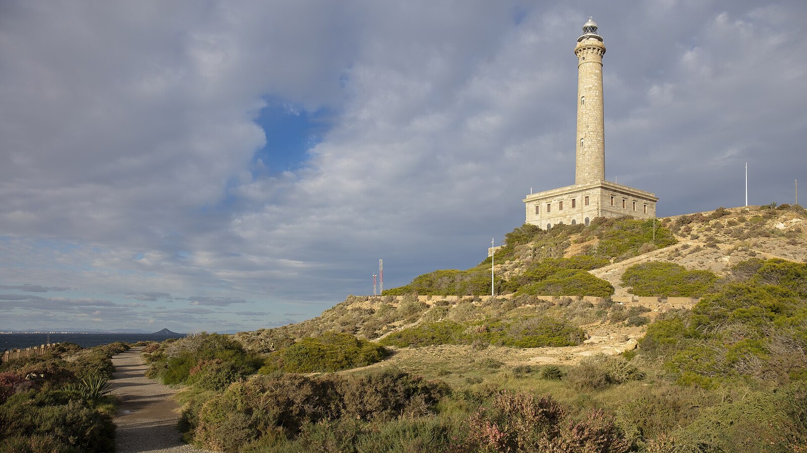 Leuchtturm von Cabo de Palos über bewachsenen Hängen, Wolkenhimmel. Wetter an der Costa Blanca und der Costa Cálida am 16.10.2026: bedeckt, 24 bis 27 Grad.