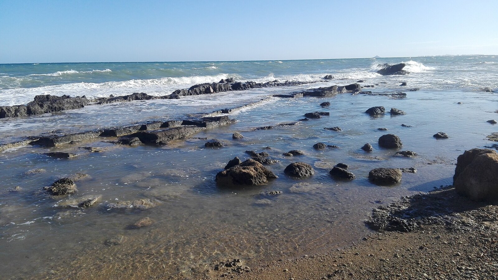 Römische Steinbrüche im flachen Wasser am Strand von Mil Palmeras, Wellen dahinter.