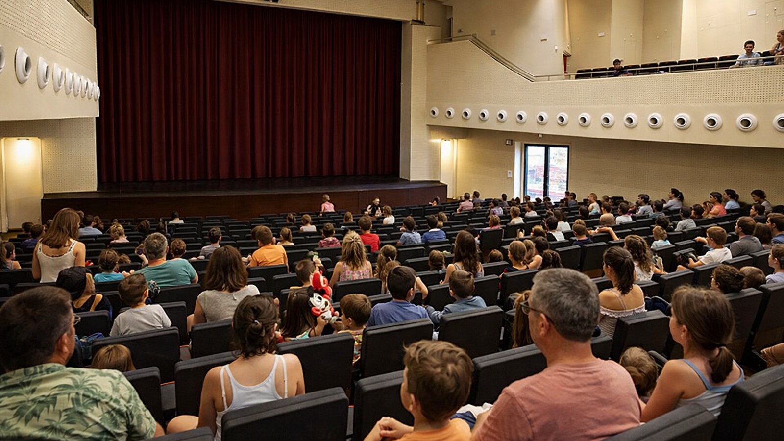 Zu sehen ist ein modernes Theaterauditorium mit beigefarbenen Wänden, dunklen gestaffelten Sitzreihen und geschlossenem roten Bühnenvorhang, in dem Familien mit Kindern kurz vor Beginn einer Vorstellung ihre Plätze einnehmen (KI-Symbolbild).