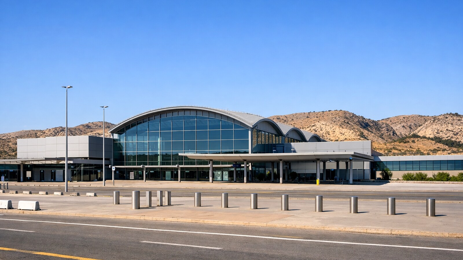 Zu sehen ist ein modernes Regionalflughafen-Terminal mit leerem Vorplatz und Straße davor, dahinter trockene mediterrane Hügel unter klarem blauem Himmel (KI-Symbolbild).