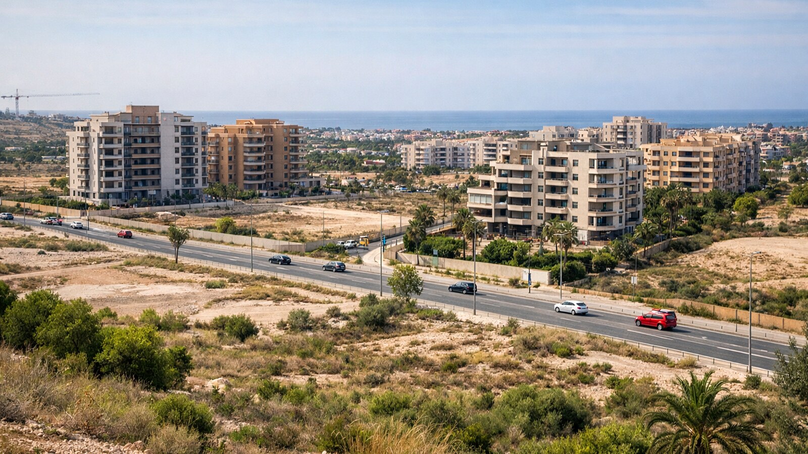 Zu sehen ist ein küstennahes Wohngebiet mit mehreren modernen mehrgeschossigen Apartmenthäusern, freien sandigen Grundstücken, einer breiten Straße mit Autos und mediterraner Vegetation im Hintergrund (KI-Symbolbild).