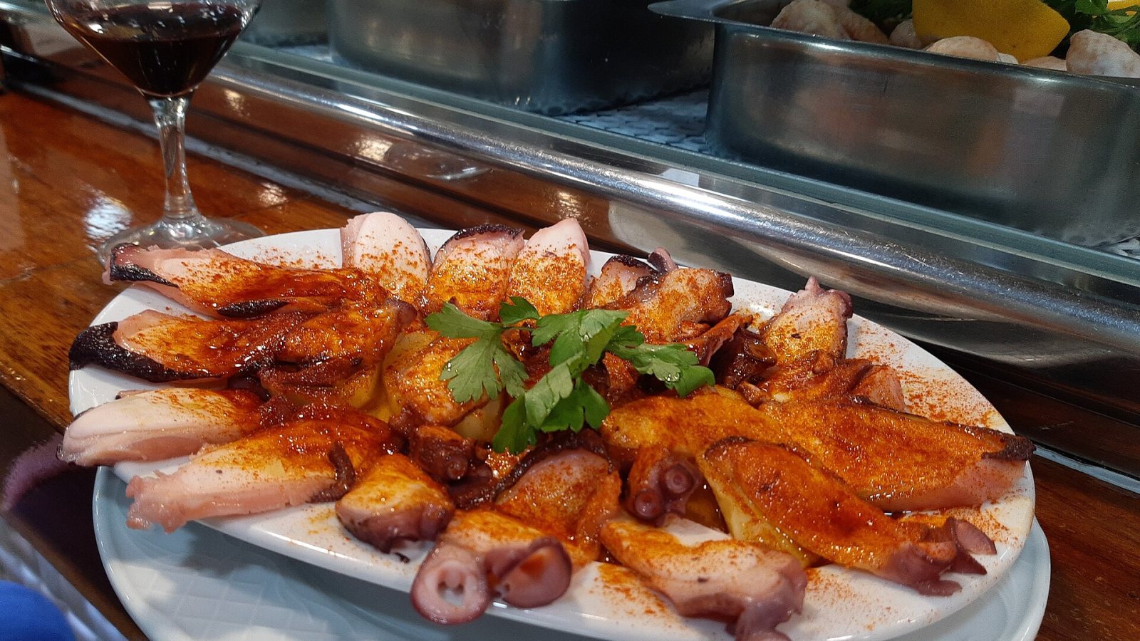 Teller mit Oktopus nach galicischer Art, mit Paprika bestreut, auf einer Bartheke, daneben ein Glas Rotwein.