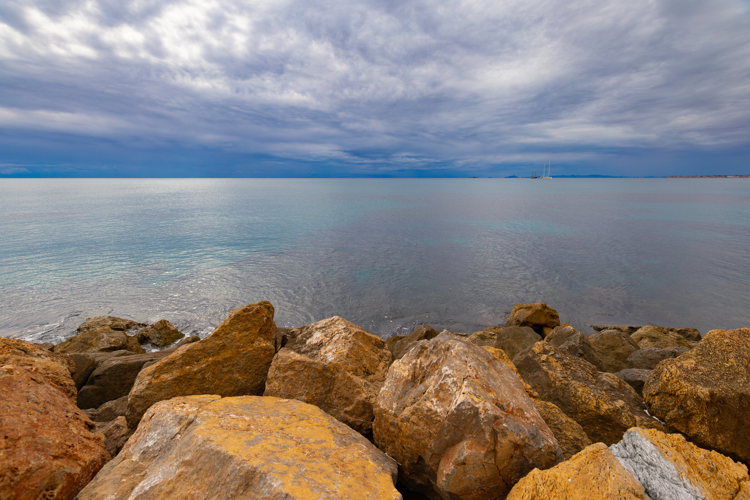 Geschlossene Wolkendecke über dem Meer, Felsblöcke im Vordergrund. Wetter an der Costa Blanca und der Costa Cálida am 19.10.2026: leichter Regen, 20 bis 23 Grad.
